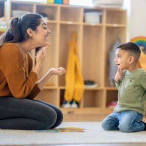 A young boy, sits on the floor with his teacher as she works through some speech therapy activities with him. They are each dressed casually and sitting on their knees as they hold their fingers on their chins and make the sounds together.
